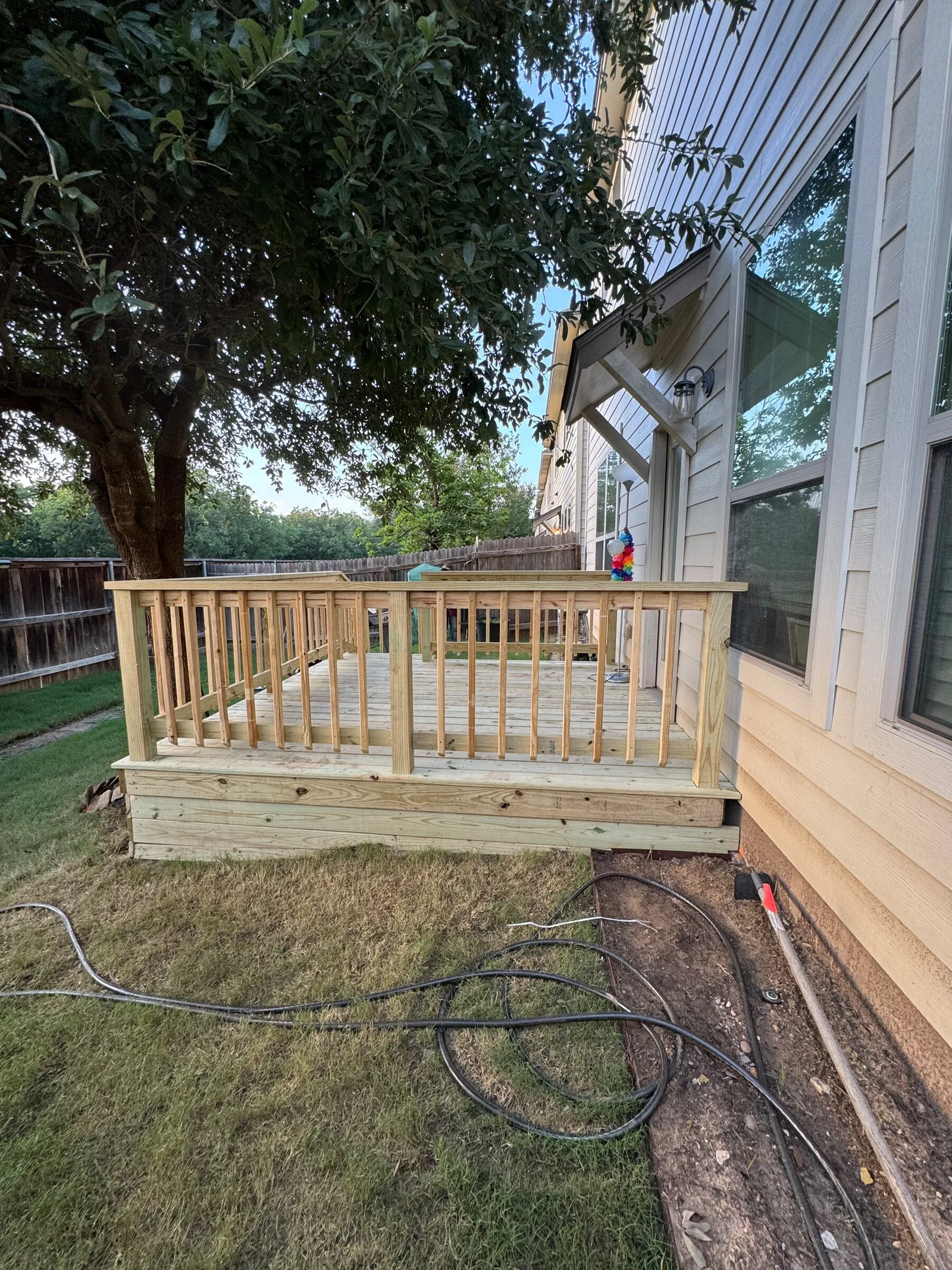 A finished wood deck with railings along the side of the house under a shade tree.
