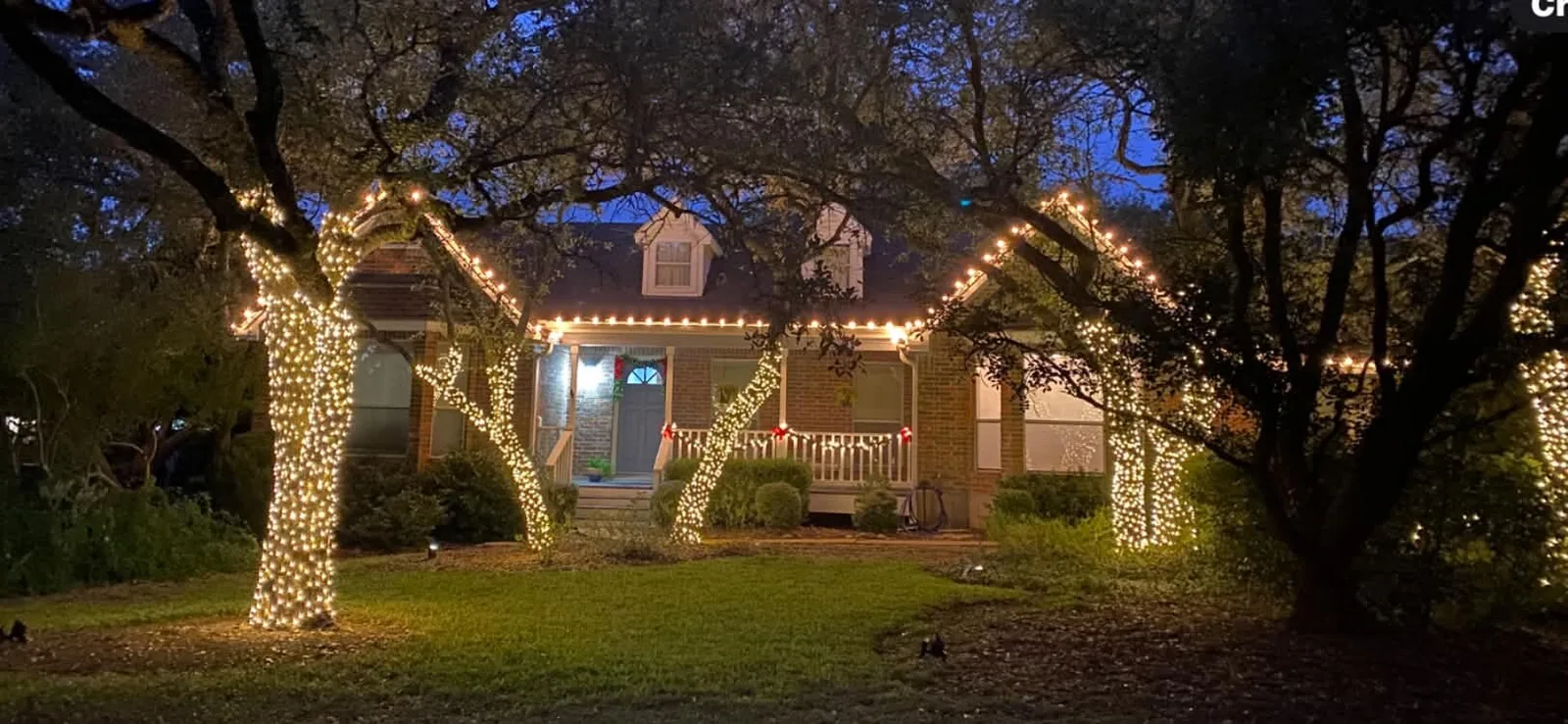 Warm white lights along a roofline and wrapped around front-yard trees at dusk.