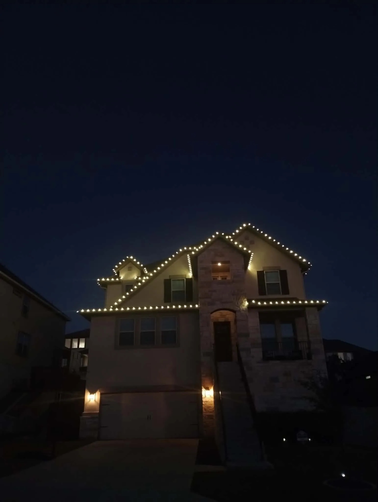 Warm white lights outlining the rooflines of a two-story stone home at night.