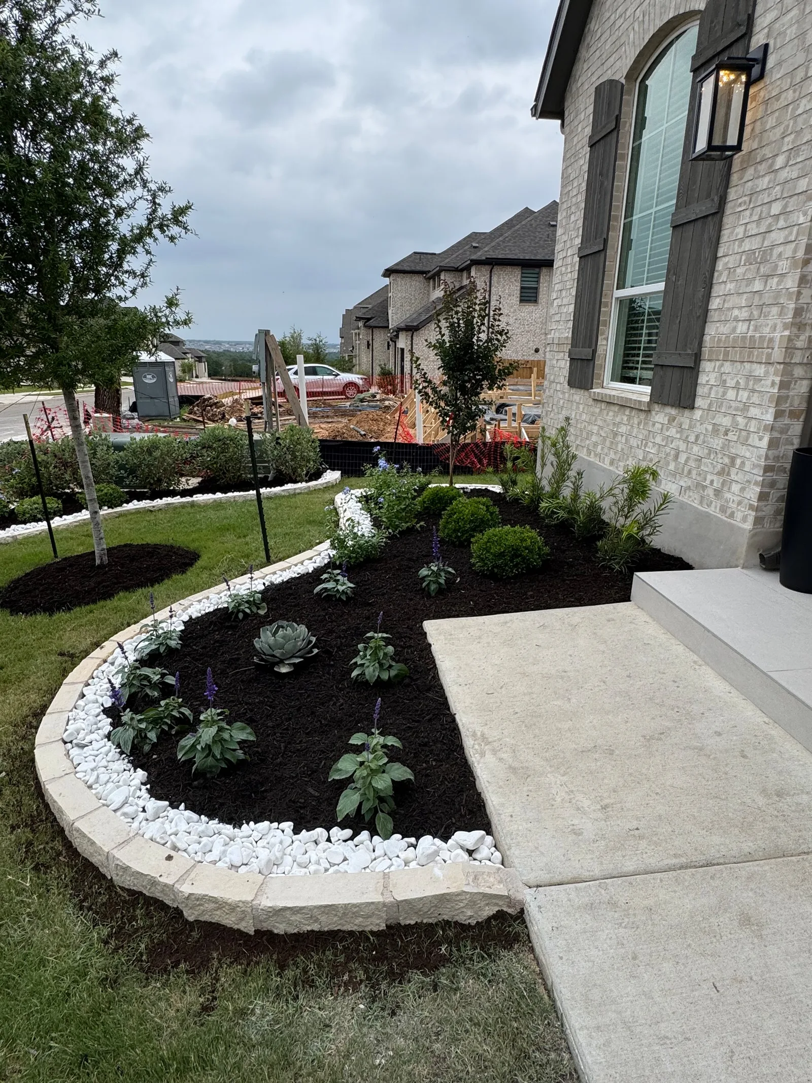 A curved planting bed with dark mulch, shrubs, white decorative rock and stone edging.