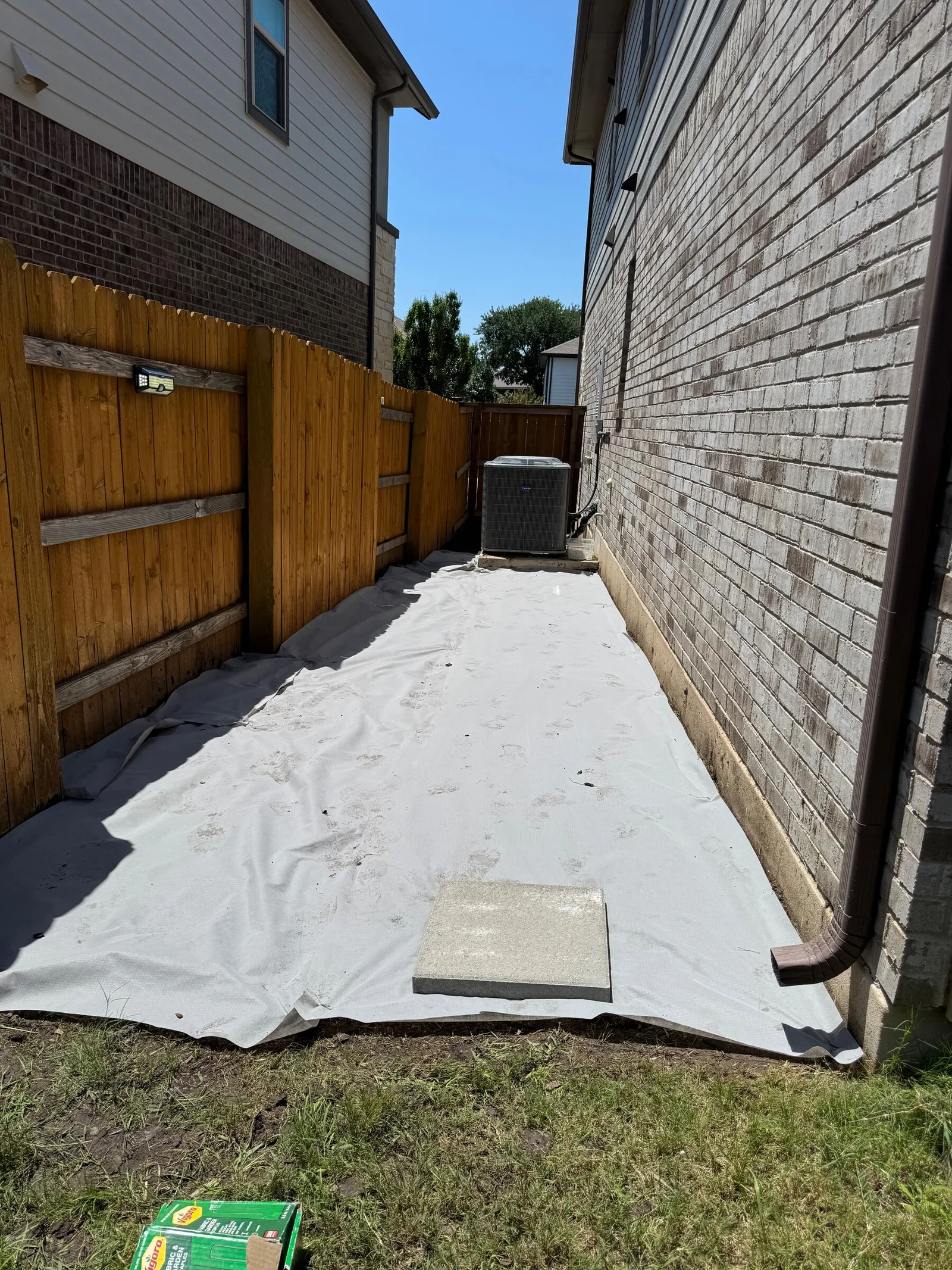 White landscape fabric covering a long side yard between a wood fence and a brick house.